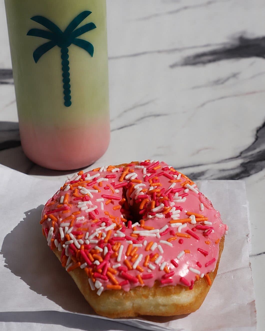 A pink sprinkle donut beside a palm-print La Costa cup on marble